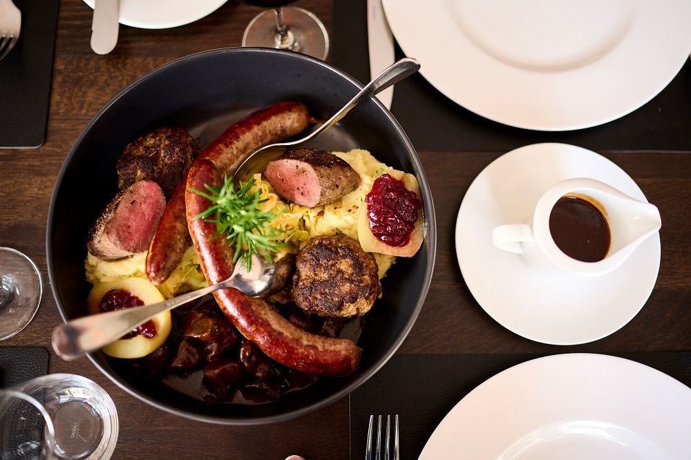 Eine schwarze Schüssel gefüllt mit Würstchen, Fleisch, Kartoffelpüree und Beerenmarmelade auf einem dunklen Holztisch mit Beilagentellern und Soße.