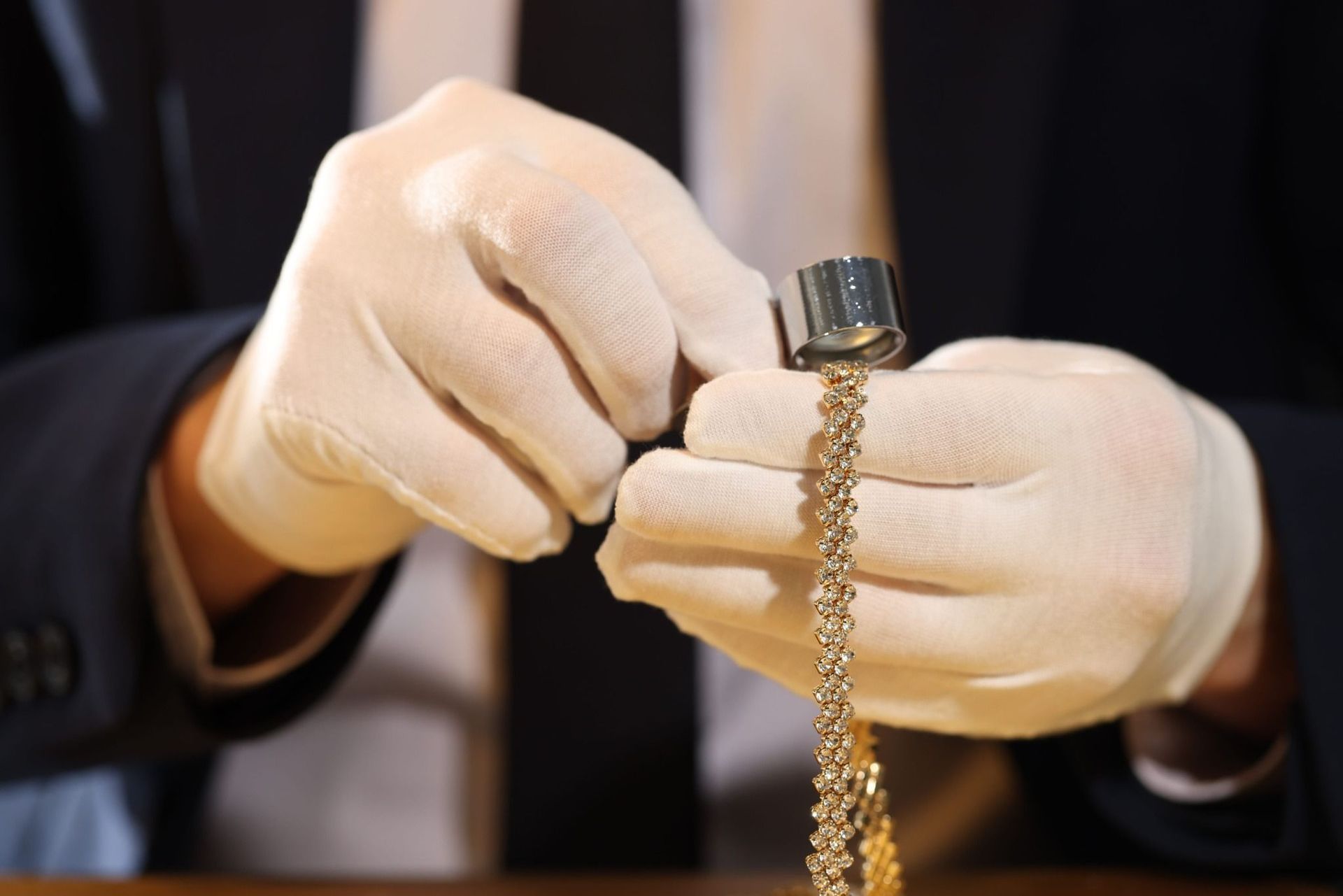 Manos enguantadas que sostienen una cadena de oro con una lupa, inspeccionando joyas sobre un fondo oscuro.