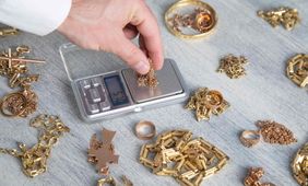 Una mano coloca joyas de oro sobre una báscula digital junto a montones de piezas de oro y un pequeño cuenco.