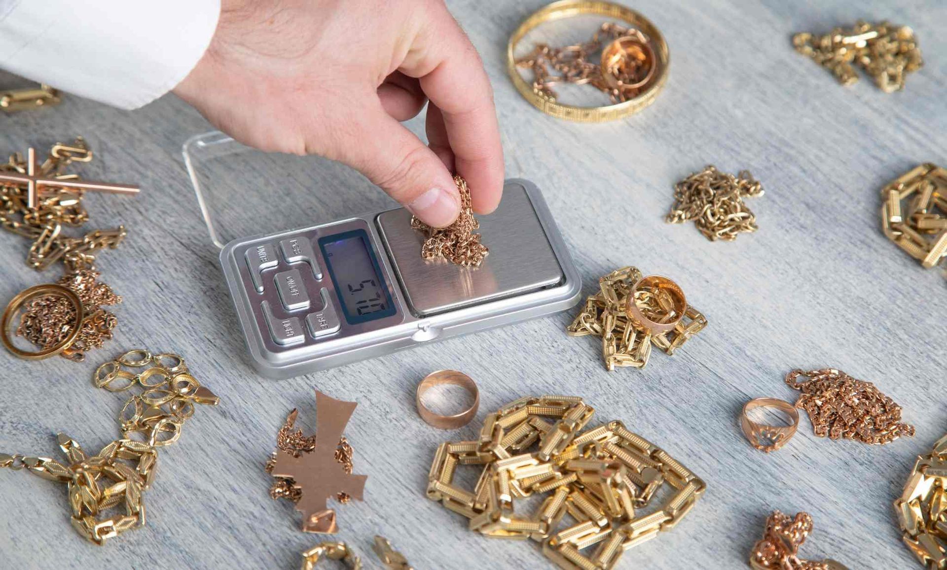 Una mano coloca joyas de oro sobre una báscula digital junto a montones de piezas de oro y un pequeño cuenco.
