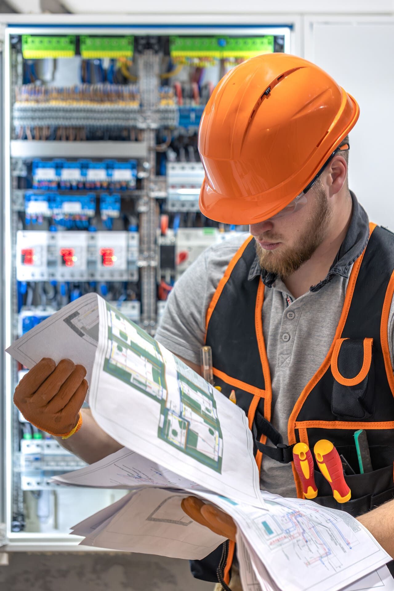 Electricista con equipo de seguridad inspeccionando planos frente a un panel eléctrico.