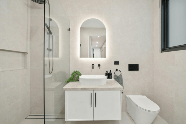 Baño moderno con paredes blancas, tocador flotante, espejo arqueado, ducha de cristal e inodoro.