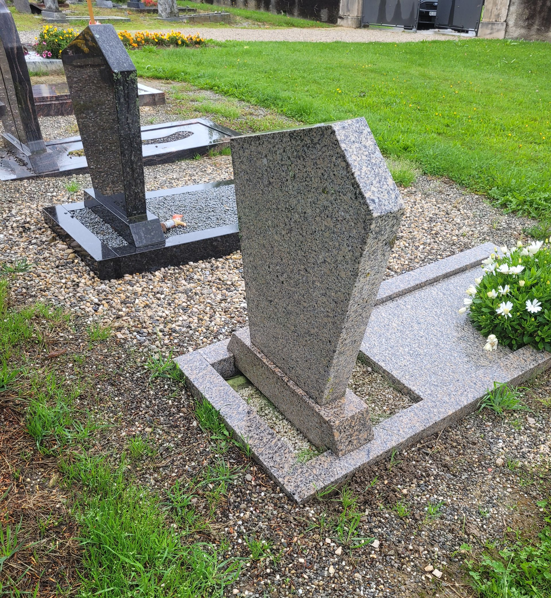 Deux pierres tombales en granit dans un cimetière, l'une droite et l'autre inclinée, à côté de gravier et d'herbe.