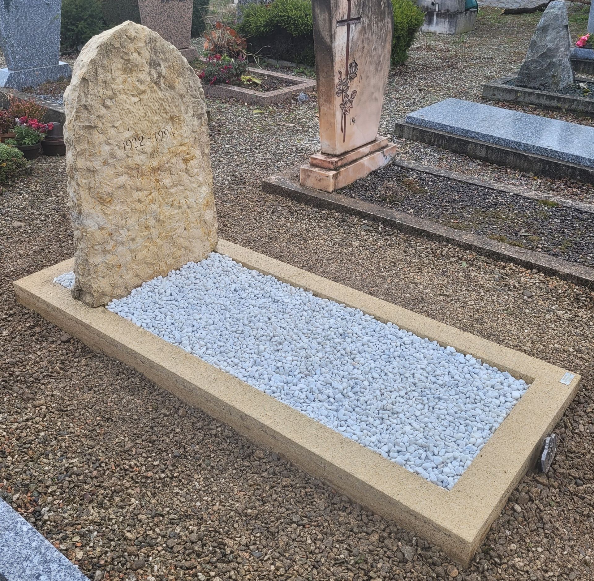 Pierre tombale commémorative en granit noir avec croix blanche et dates, posée sur un terrain gravillonné.