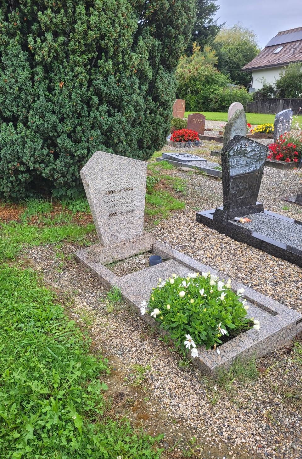 Deux pierres tombales dans un cimetière-jardin, l'une blanche et l'autre sombre, entourées de fleurs et de verdure.