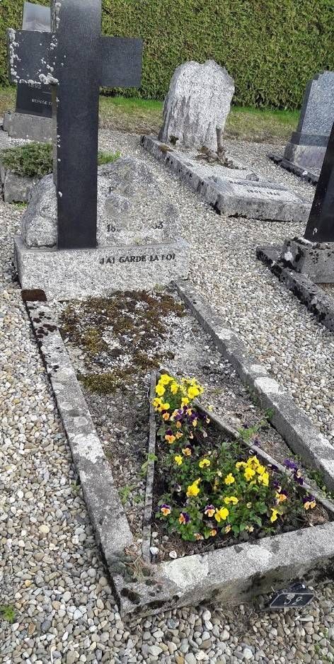 Pierre tombale dans un cimetière, à côté d'une tombe fleurie et de pierres tombales recouvertes de mousse.