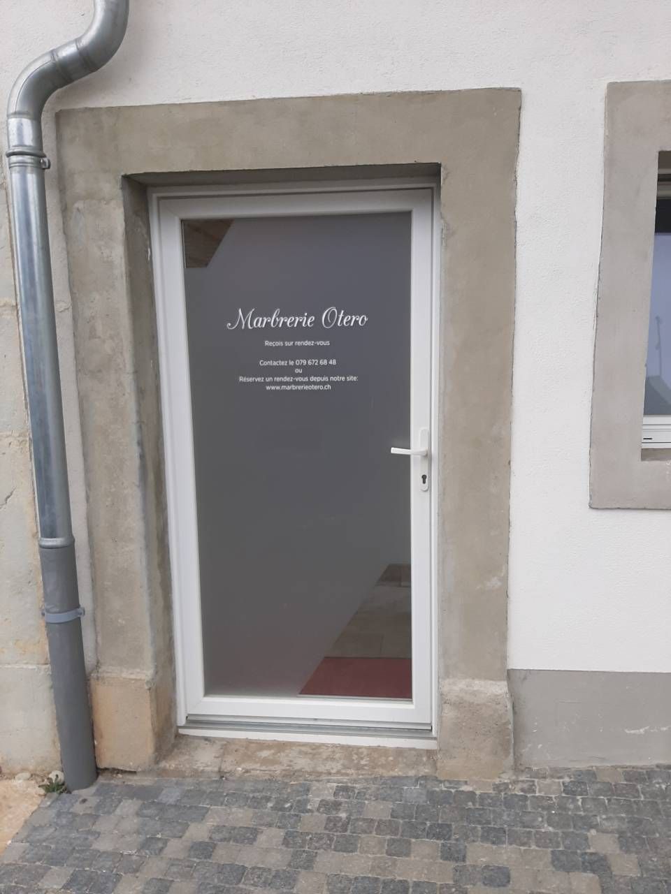 Porte vitrée avec inscription blanche sur un mur beige et trottoir pavé à l'extérieur