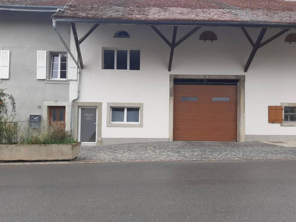 Façade blanche d'un bâtiment avec une porte de garage en bois, de petites fenêtres et une allée pavée en bord de route.