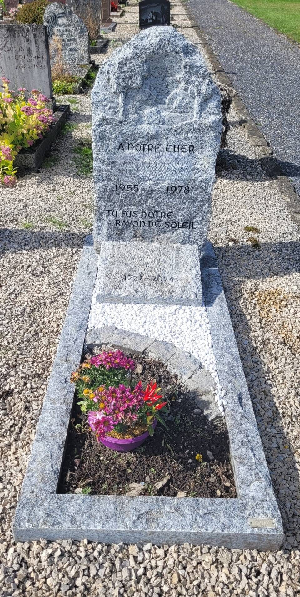 Pierre tombale avec un petit pot de fleurs dans un cimetière, encadrée par des allées de gravier.