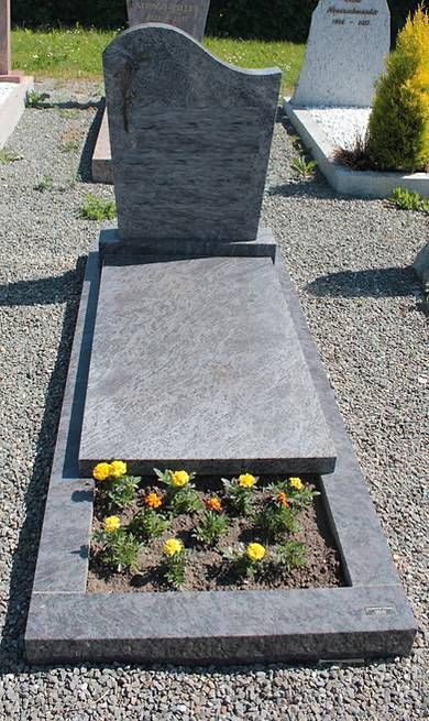 Pierre tombale grise avec parterre de fleurs dans un cimetière, entourée de gravier et d'autres pierres tombales.