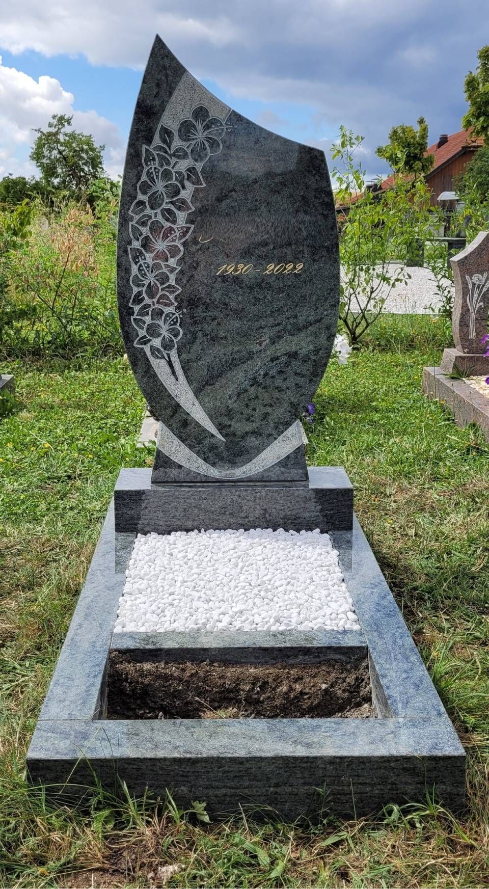 Pierre tombale commémorative en granit dans un cimetière herbeux, avec une pierre sombre incurvée sur une base blanche.