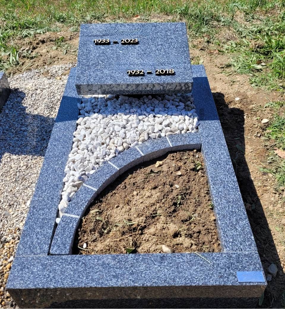 Pierre tombale en granit bleu avec une découpe incurvée, pierres blanches et terre apparente dans un cimetière
