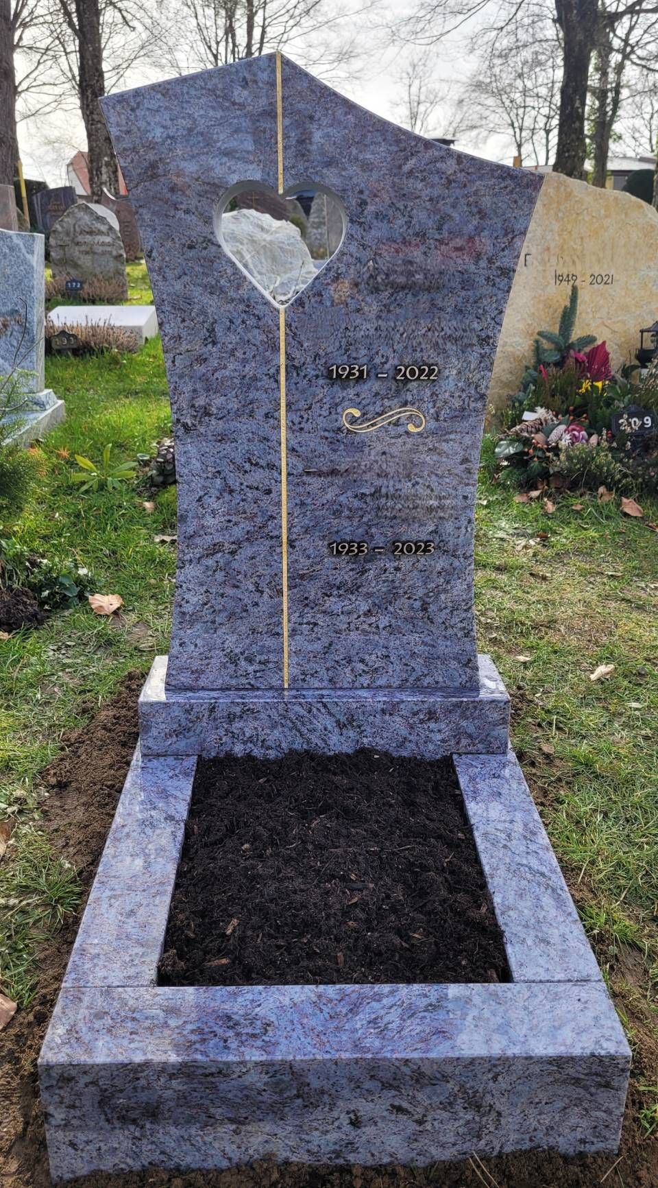 Pierre tombale en granit gris avec une découpe en forme de cœur dans un cimetière, encadrée de fleurs et d'arbres