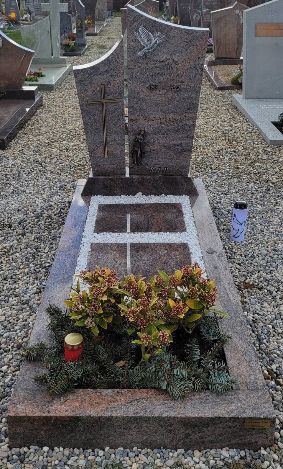 Pierre tombale avec une croix blanche dans un cimetière, bordée de fleurs et de gravier.