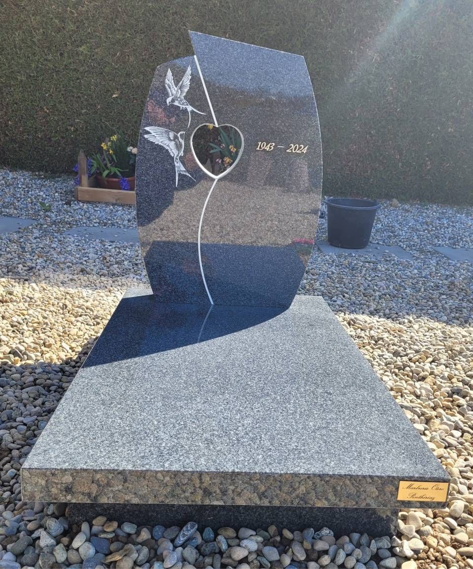 Monument abstrait en pierre noire avec une découpe en forme de cœur sur un socle de gravier dans un jardin.