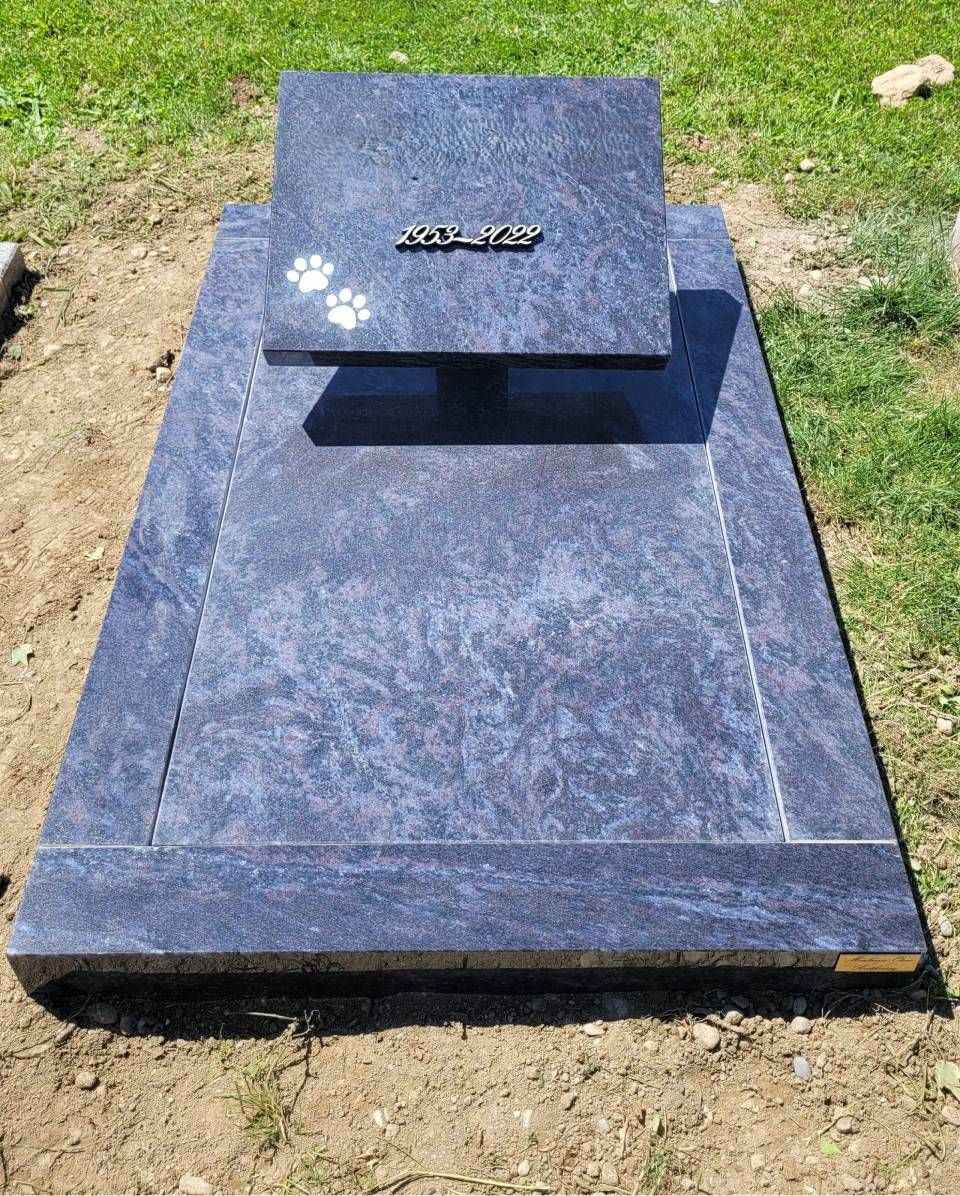 Pierre tombale en granit bleu dans un cimetière herbeux, avec une stèle surélevée.