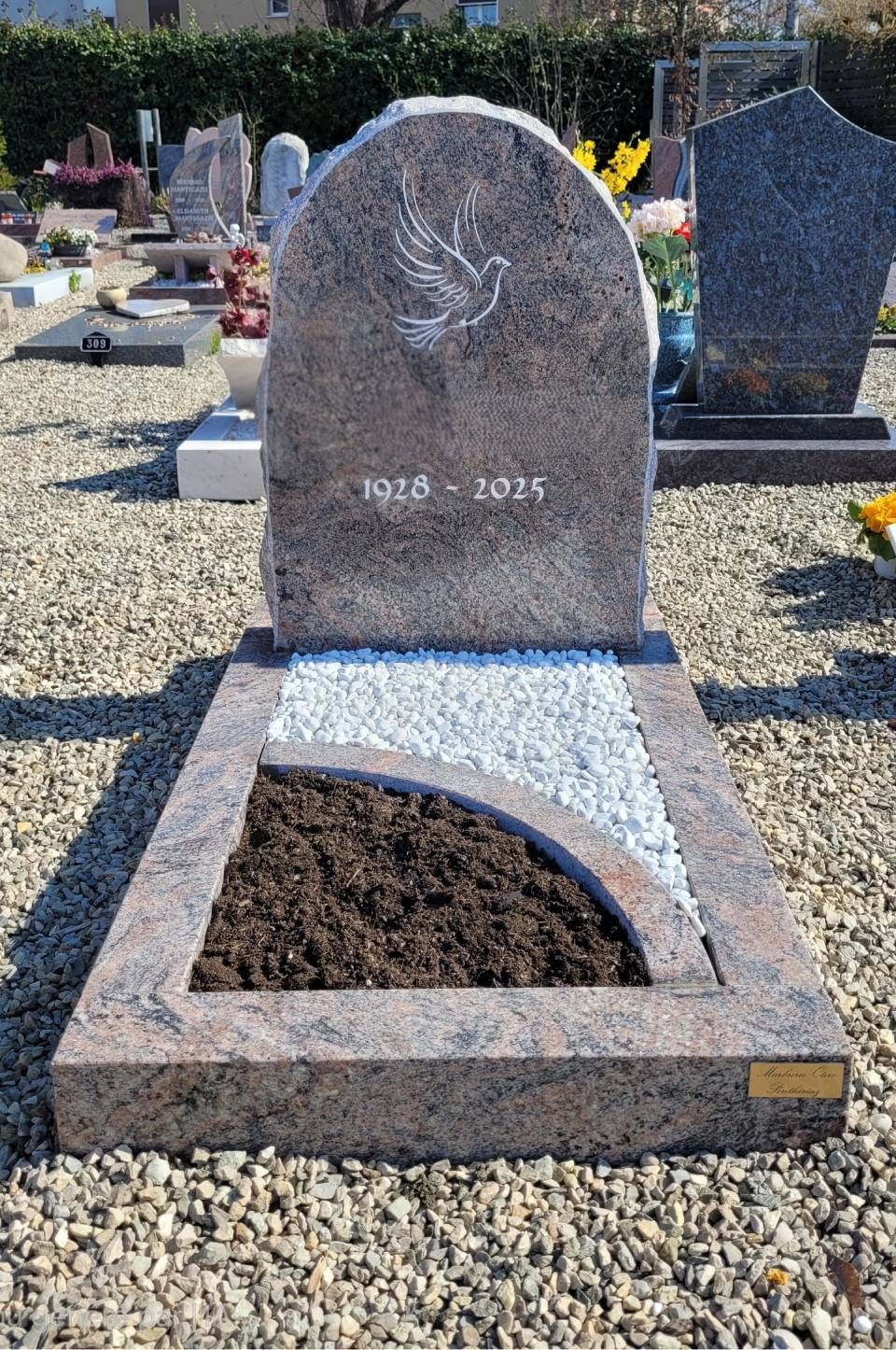Pierre tombale grise avec un ange gravé dans un cimetière, située dans une parcelle remplie de cailloux et de terre sombre.