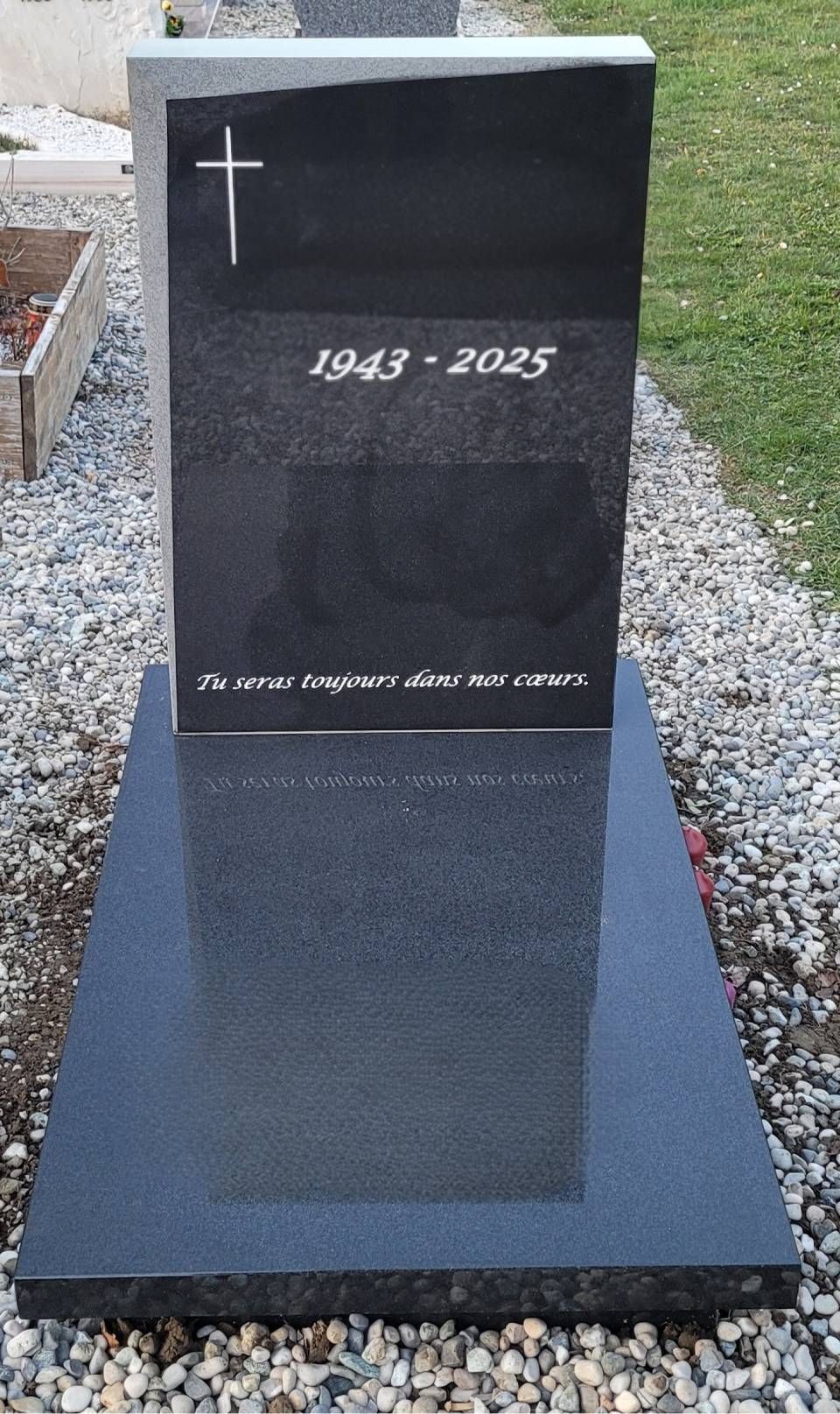 Pierre tombale commémorative en granit noir avec croix blanche et dates, posée sur un terrain gravillonné.