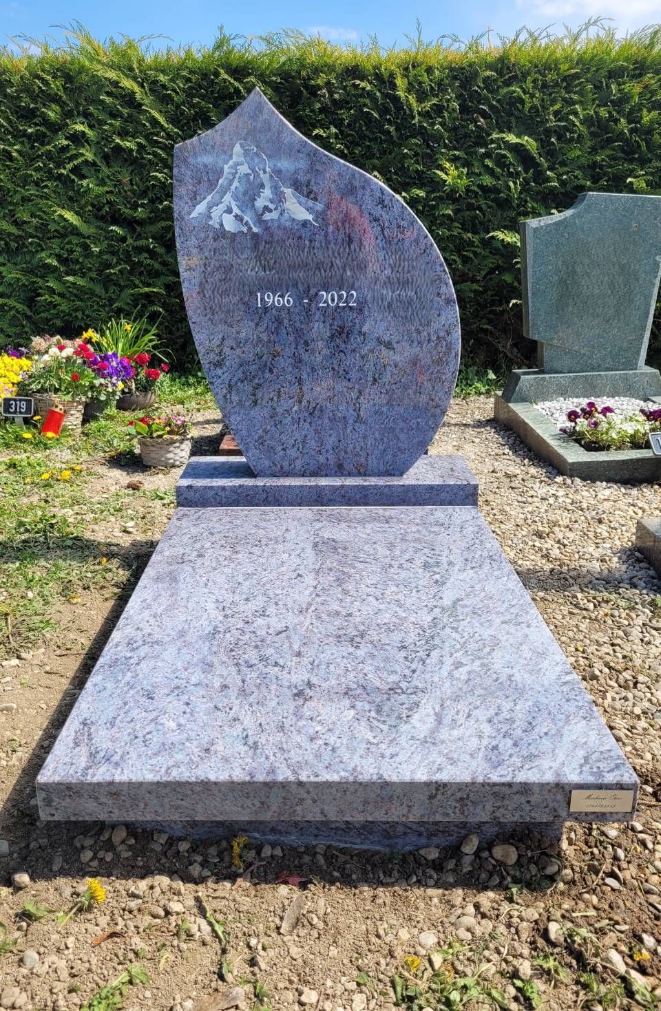 Pierre tombale en granit à base inclinée dans un cimetière, bordée de fleurs et d'une haie verte