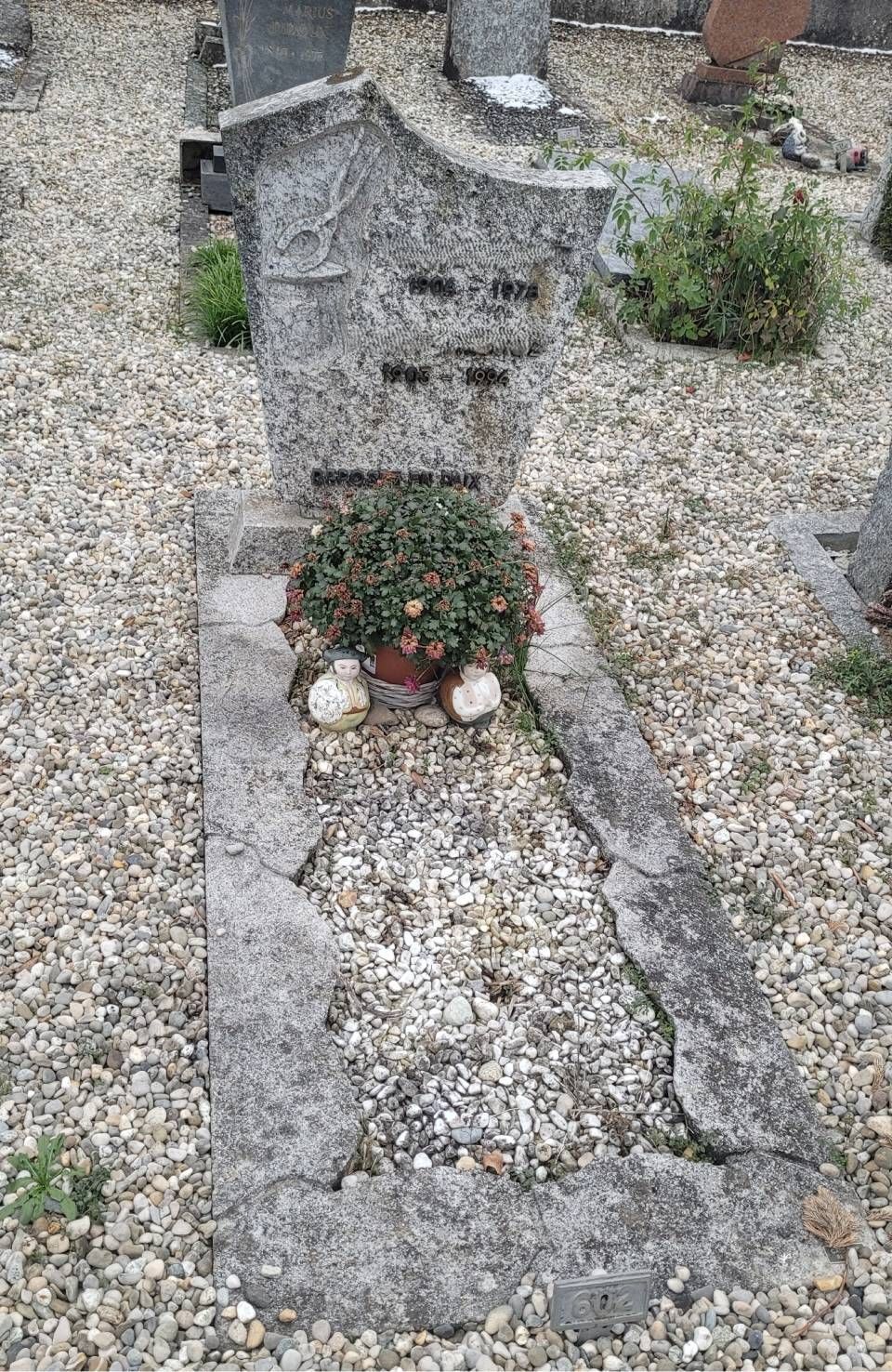 Pierre tombale dans une parcelle de cimetière gravillonnée, ornée de petites fleurs à sa base