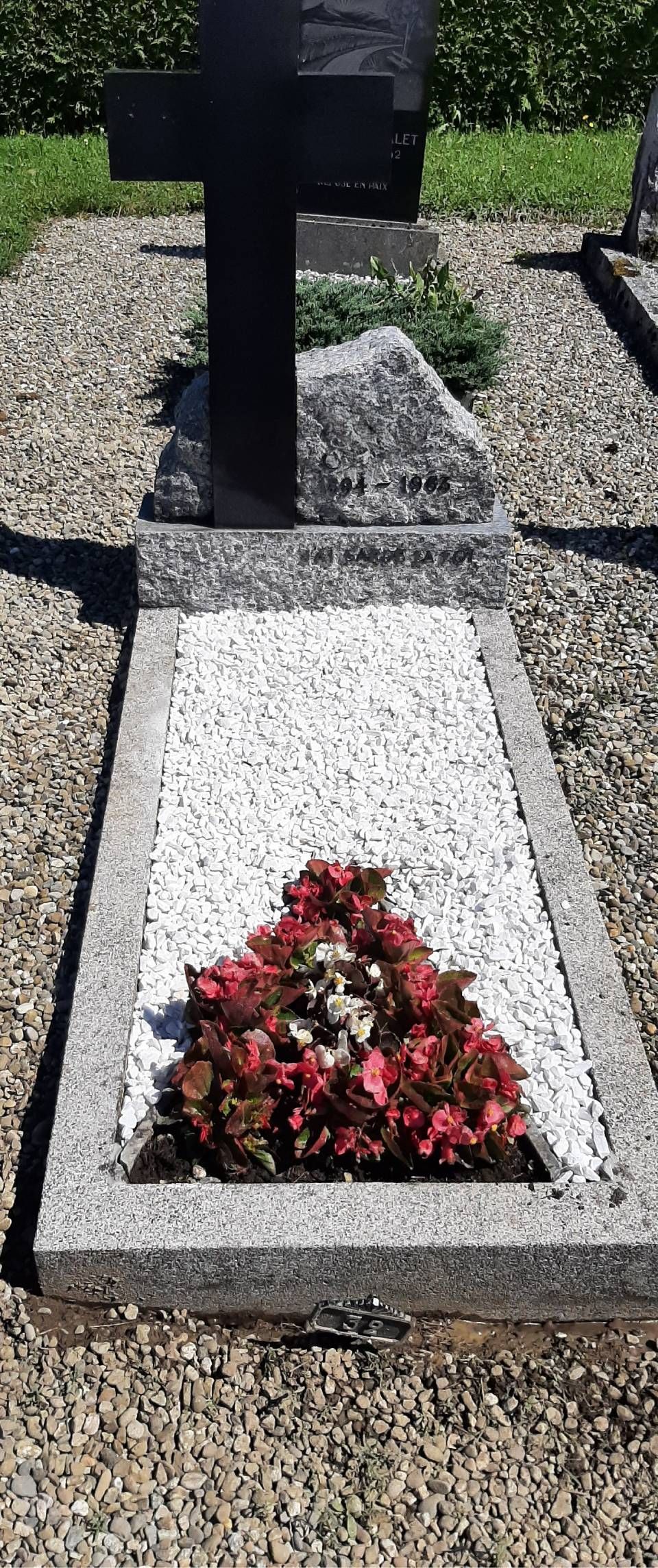 Tombe en gravier blanc ornée d'un petit arrangement de fleurs rouges et d'un monument funéraire sur poteau noir.