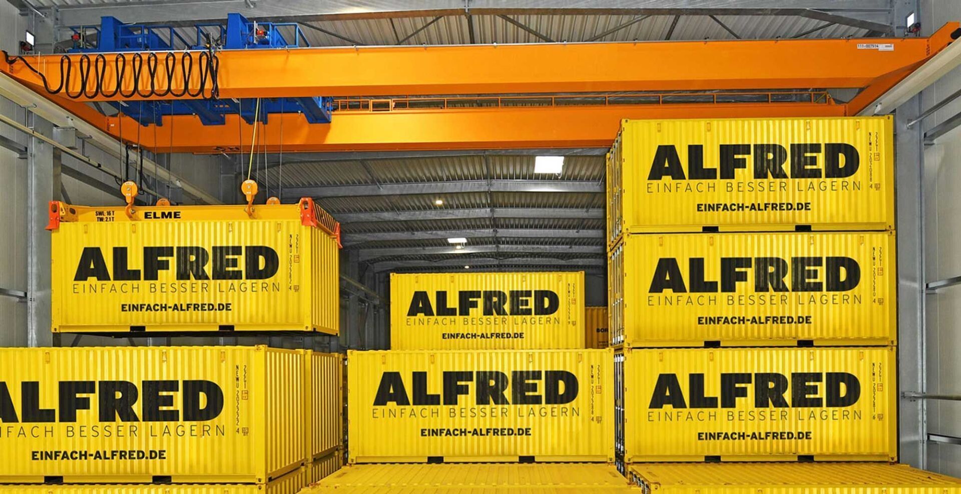 Brückenkran hebt einen gelben Container mit der Aufschrift „ALFRED“ in einer Lagerhalle an.