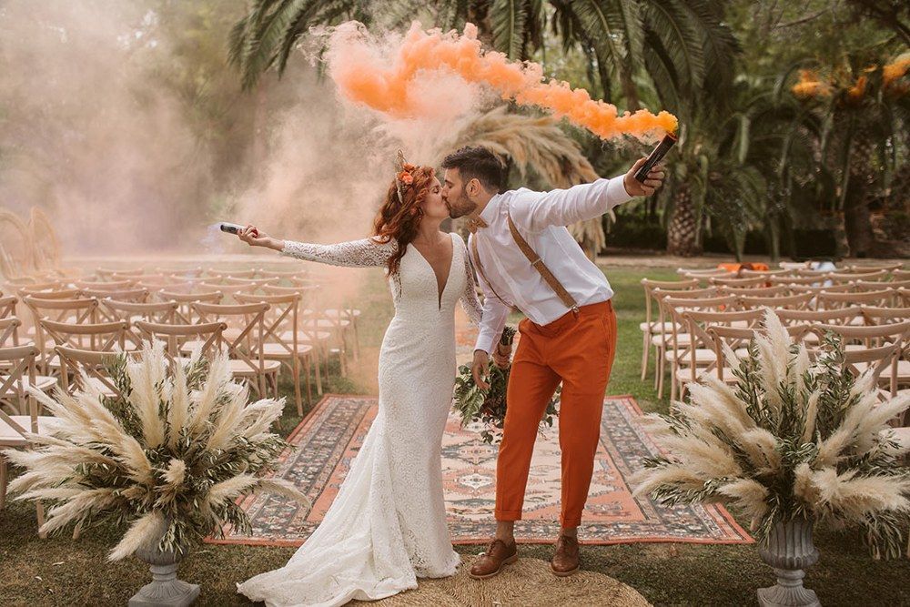 Una pareja se da un beso con polvos de colores en una celebración al aire libre entre palmeras y alfombras.