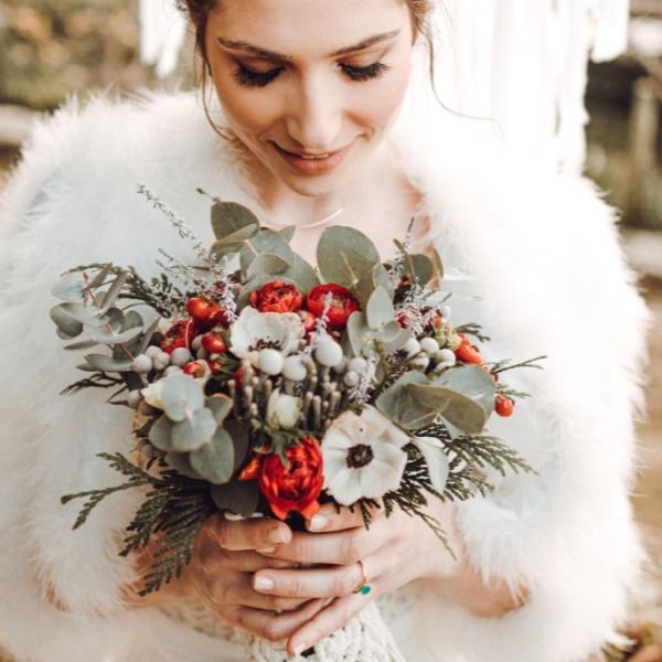Mujer sosteniendo un ramo de invierno con bayas rojas, flores blancas y follaje, vistiendo una manta blanca y esponjosa.
