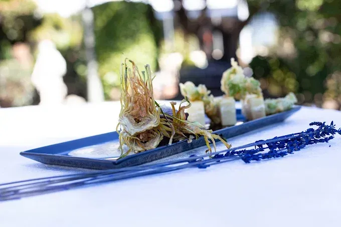 Aperitivo en plato azul, alcachofa frita, verduras, mantel blanco y escenario exterior difuminado.