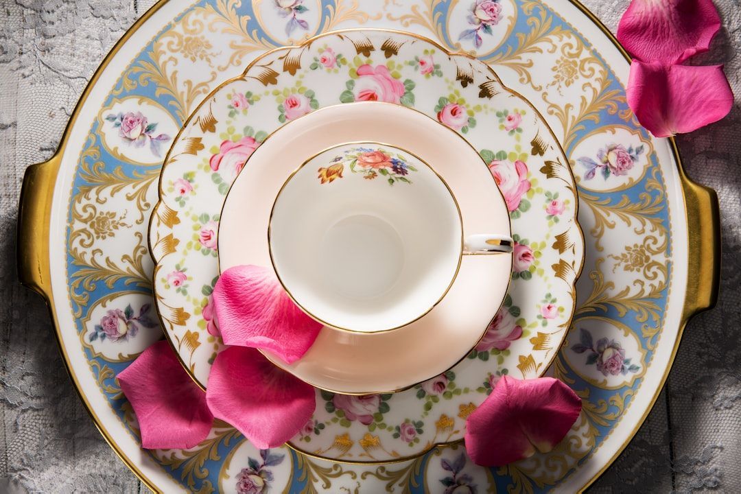 Taza y platillo con motivos florales sobre un plato ornamentado con borde dorado y pétalos de rosa rosa.