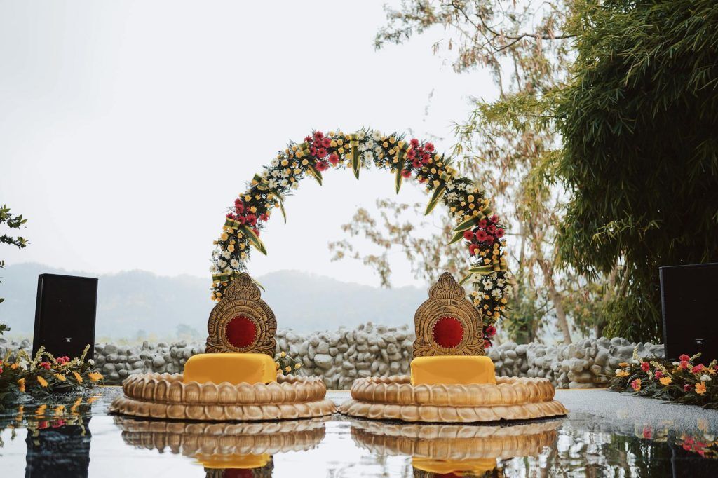 Escenario floral para bodas al aire libre con dos sillas doradas decoradas y un arco de flores junto al agua.