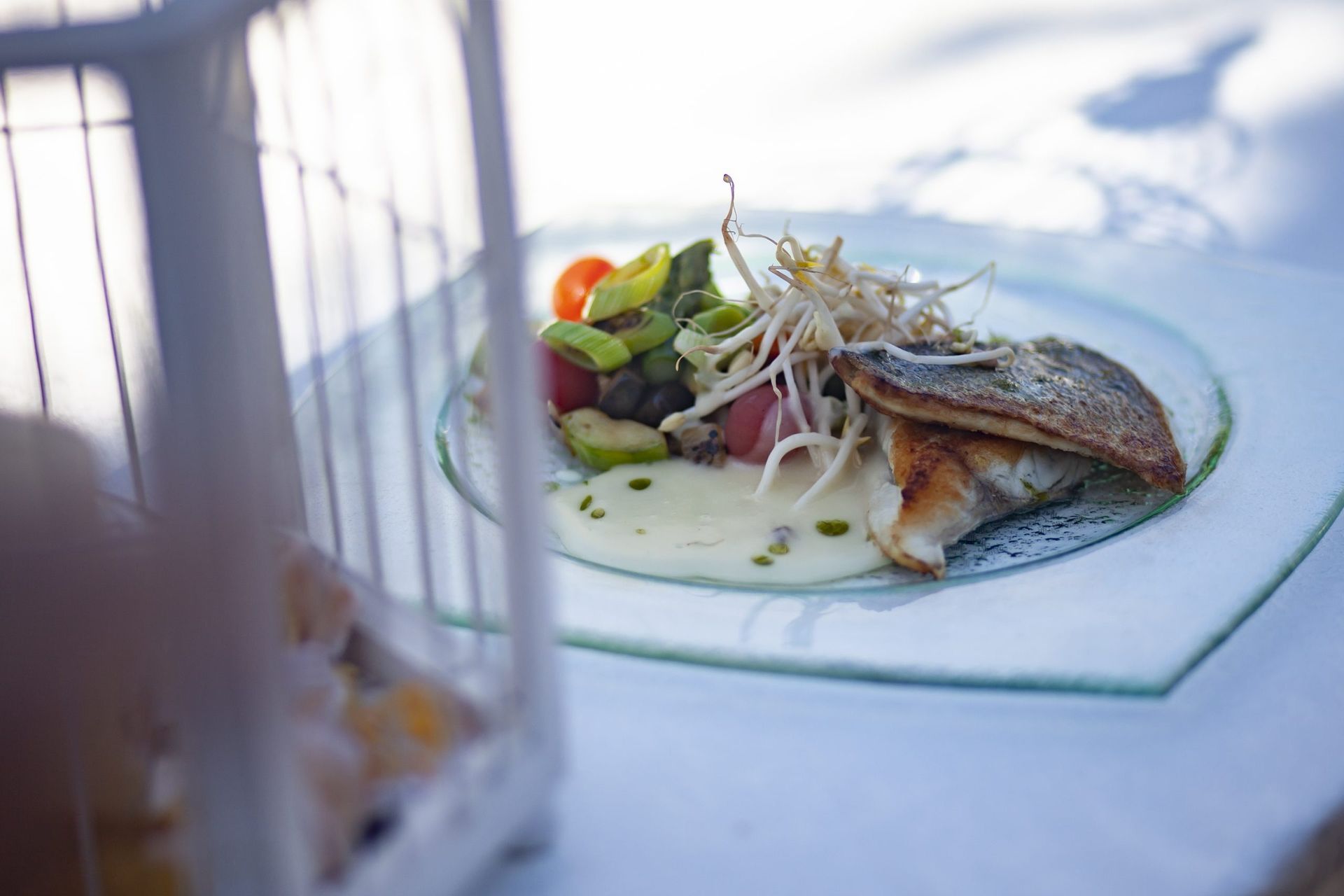 Plato principal de pescado en un plato de cristal con verduras, brotes y salsa, sobre un mantel blanco, primer plano borroso.