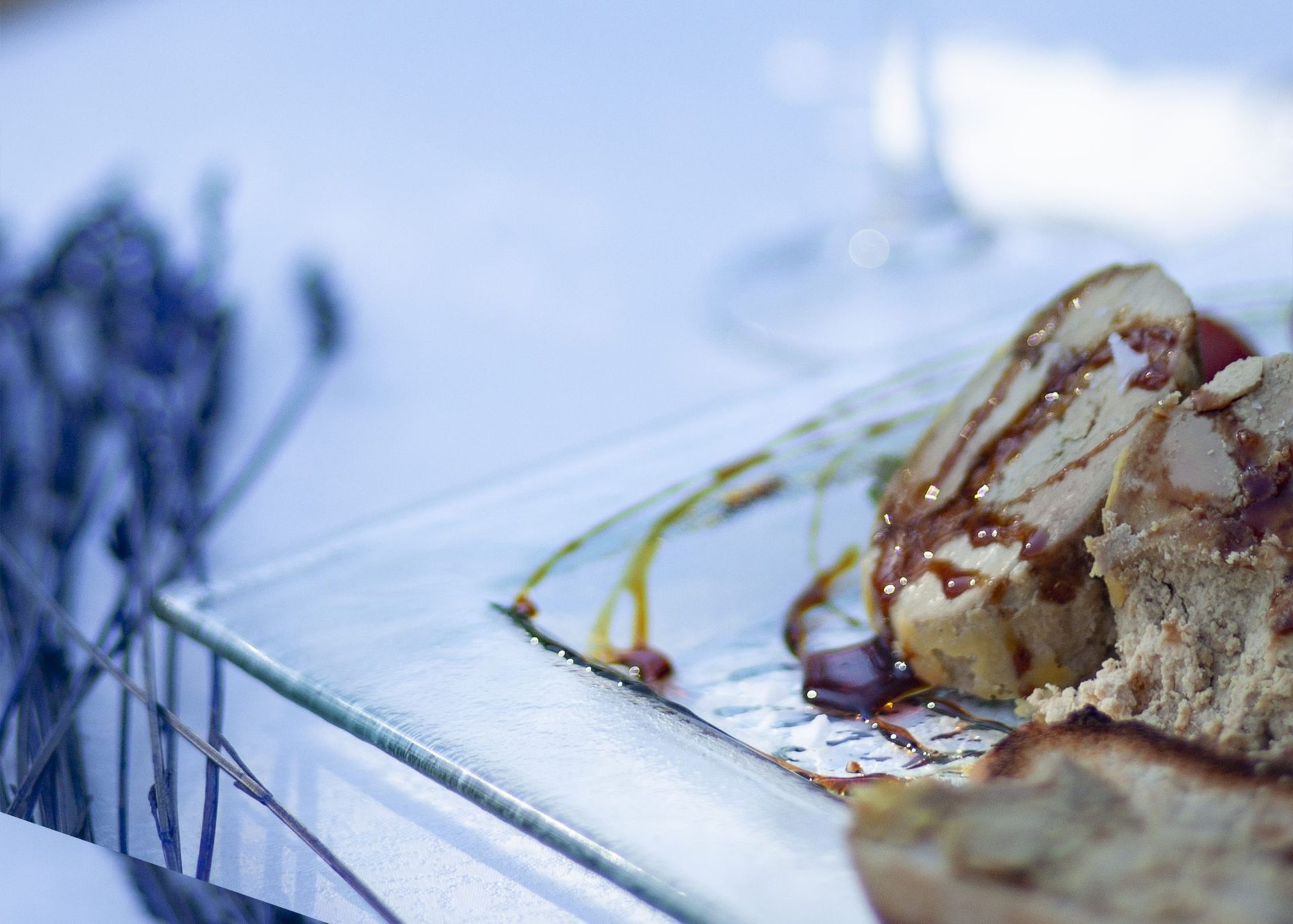 Primer plano de comida en un plato transparente, adornado con salsa y pan, junto a lavanda y un mantel blanco.