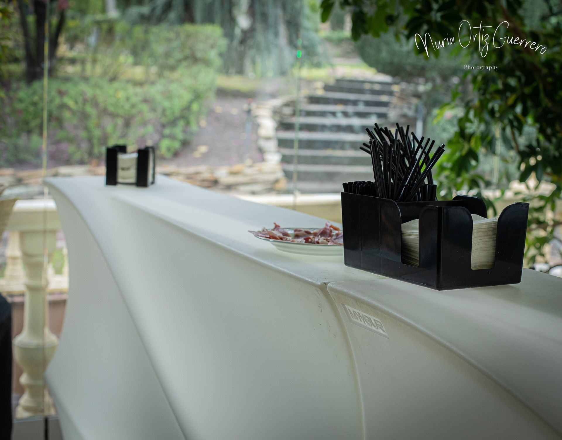 Barra blanca con accesorios negros: pajitas y servilletas. Fondo exterior borroso con escaleras.