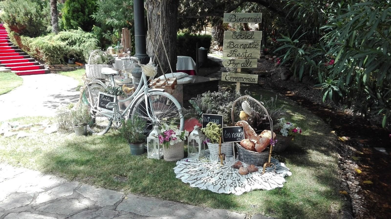 Exhibición decorativa con una bicicleta blanca, flores, carteles y un osito de peluche en un entorno de jardín.