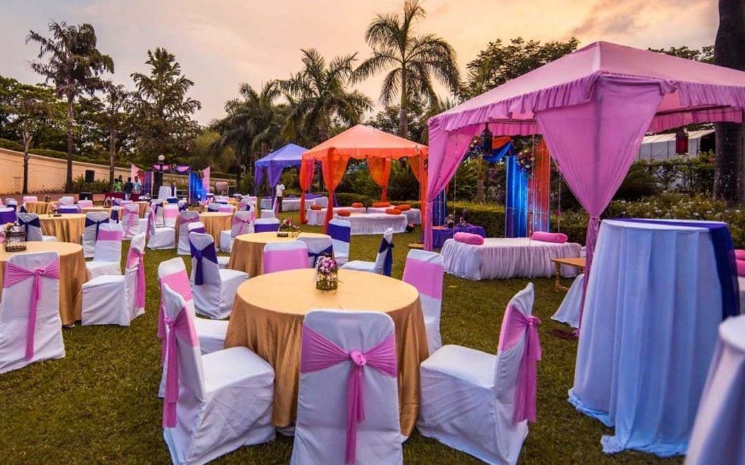 Evento al aire libre con mesas y sillas decoradas en tonos rosa, dorado y azul; bajo toldos de colores.