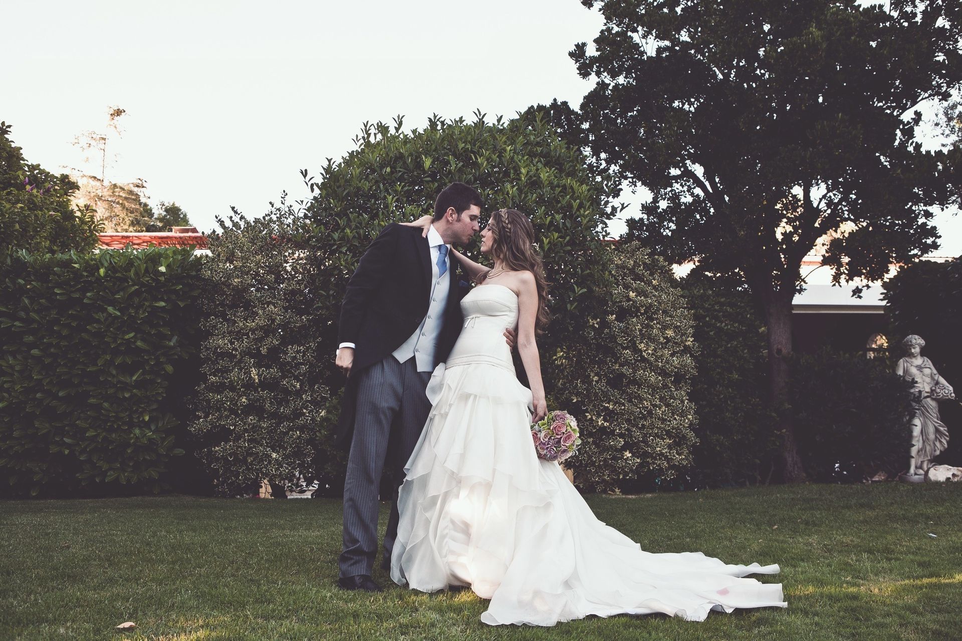 Pareja abrazándose al aire libre; novia con vestido blanco, novio con traje, entorno de jardín.