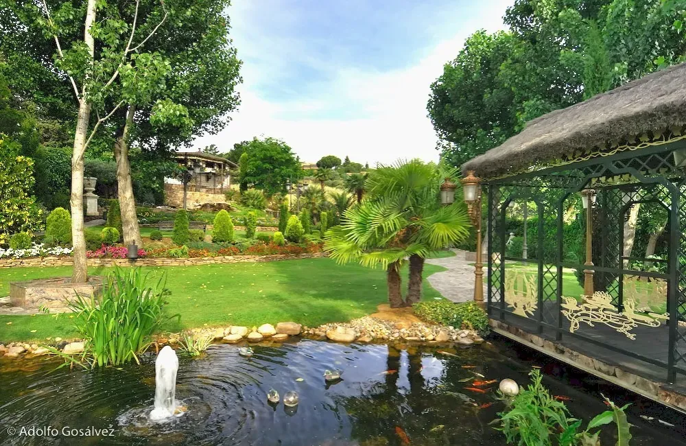 Jardín exuberante con estanque y fuente. Hay un cenador a la derecha. Césped verde, árboles y flores de colores.