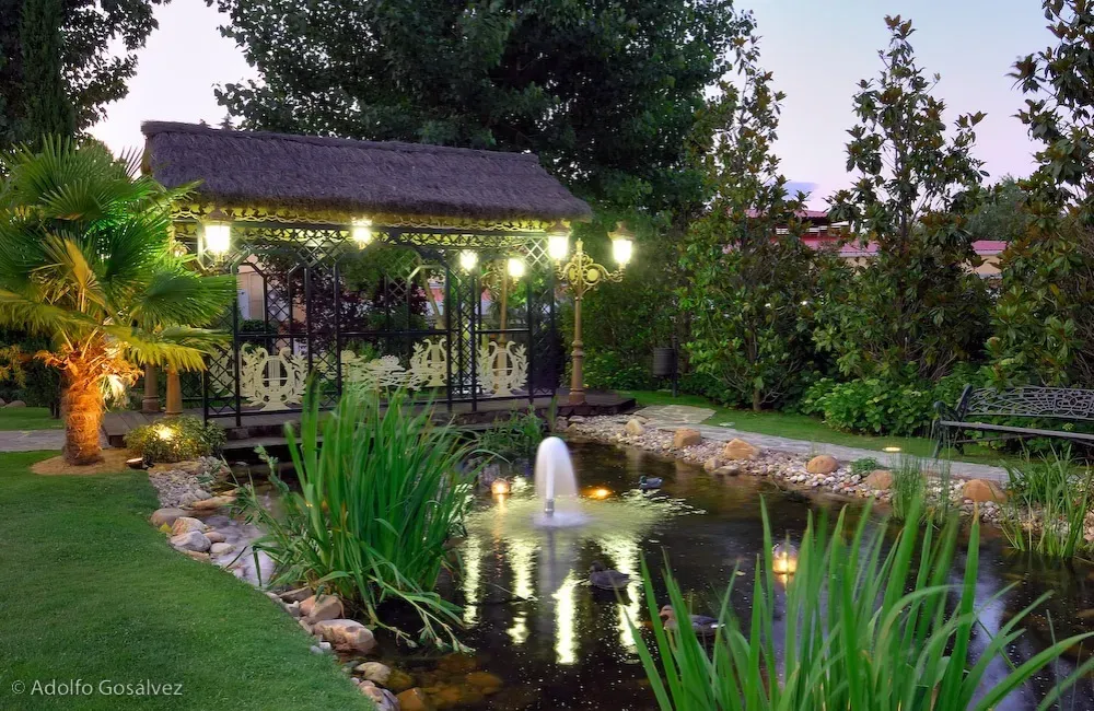 Estanque con fuente, cenador decorativo y exuberante vegetación en un entorno de jardín, iluminado por luces suaves.