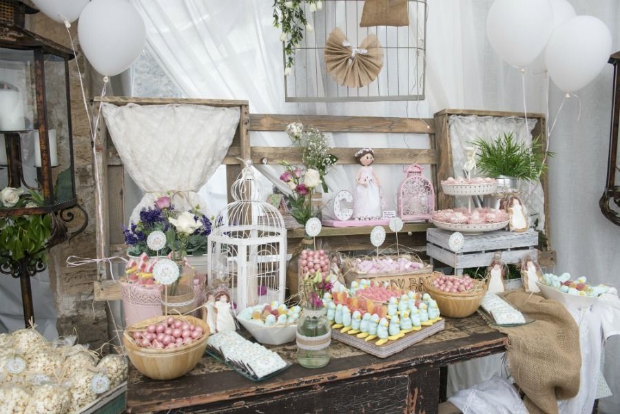 Mesa de postres decorada con golosinas blancas, rosas, flores, globos y elementos rústicos de madera.