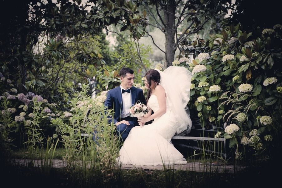 Los novios están sentados en un banco en un jardín, rodeados de flores. El novio lleva traje; la novia, vestido blanco y velo.