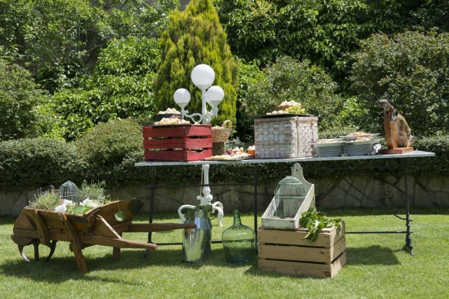 Exposición de alimentos al aire libre sobre mesa y cajas, incluyendo carretilla y entorno de jardín.