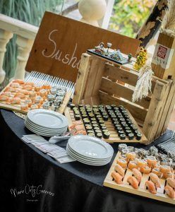 Buffet de sushi en una mesa negra con un cartel de madera y platos apilados, al aire libre.