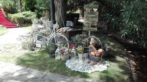 Bicicleta vintage blanca con decoraciones florales, cestas y un cartel en un jardín al aire libre.