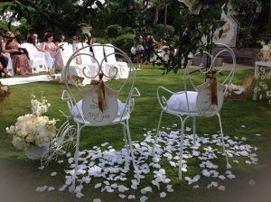 Dos sillas decoradas para una ceremonia de boda en el césped, cubiertas de pétalos blancos. Los invitados al fondo.