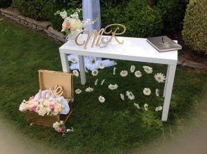 Mesa de recepción de boda sobre césped con flores, iniciales y libro de visitas.
