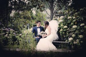 Los novios están sentados en un banco de un jardín, rodeados de flores. El novio lleva traje y la novia sostiene un ramo.