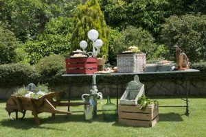 Exhibición de comida al aire libre sobre una mesa y cajas rústicas, con vegetación y decoración de vidrio en un jardín soleado.