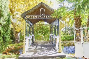 Puente de madera decorativo sobre un pequeño estanque con árboles y faroles; entorno luminoso y soleado.