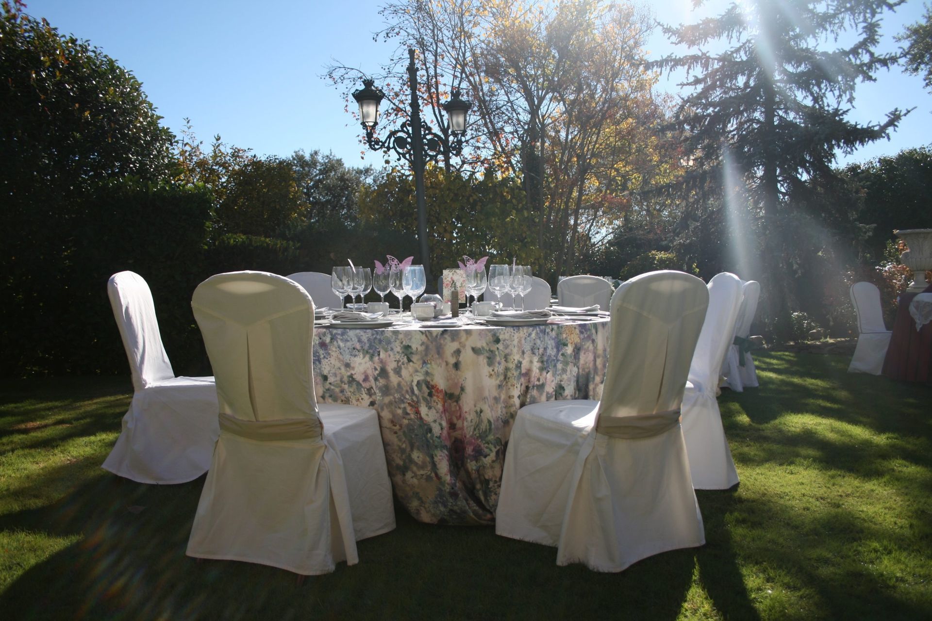 Mesa redonda para evento al aire libre, rodeada de sillas, mantel floral y ambiente de jardín soleado.
